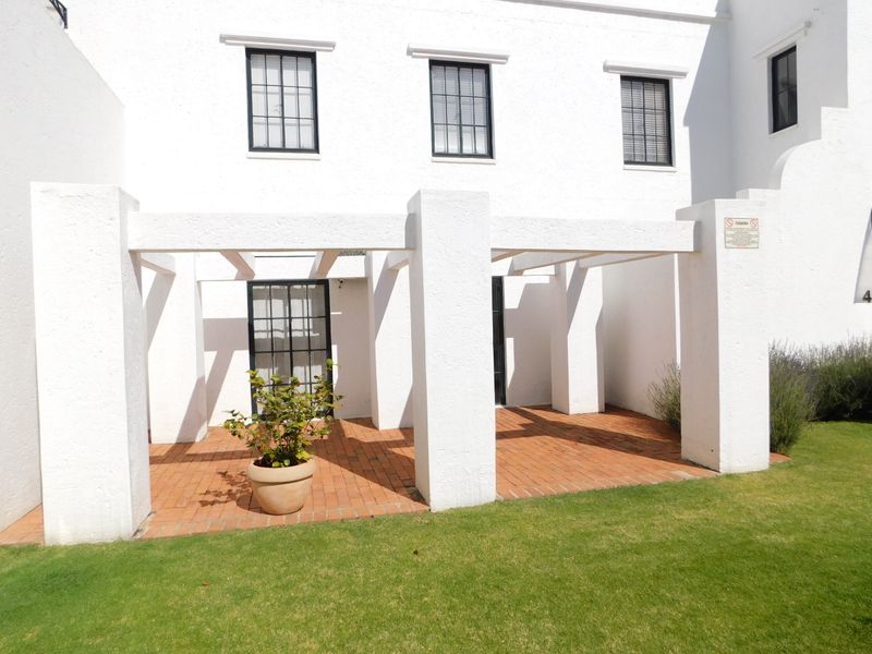 A covered patio area featuring brick paving and a structural pergola, adjacent to a well-maintained lawn and white building facade.