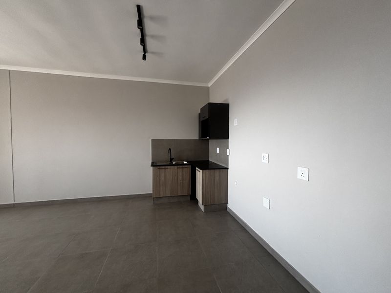 A modern, minimalist open-plan living area featuring a compact kitchenette with dark cabinetry, granite countertops, and sleek track lighting.