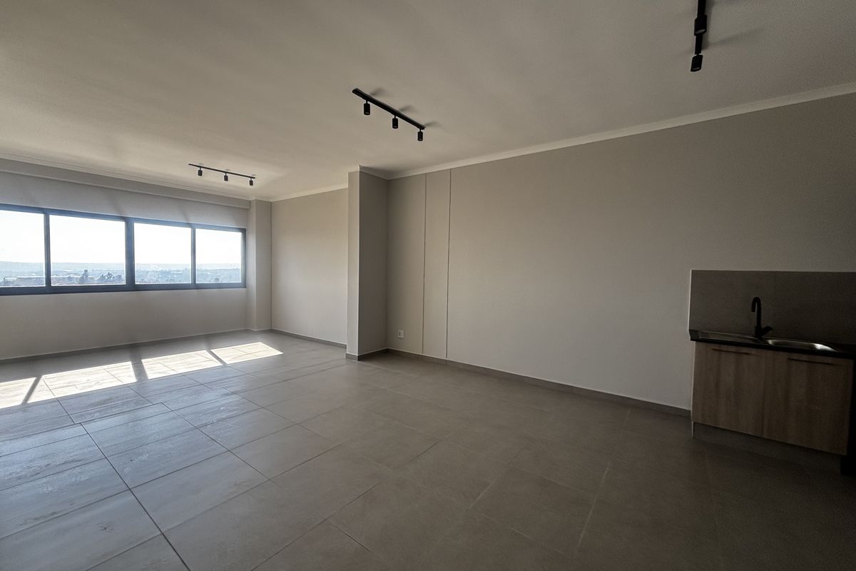Spacious open-plan living area featuring modern tiled flooring, track lighting, and a compact kitchenette with a sink, offering bright natural light.