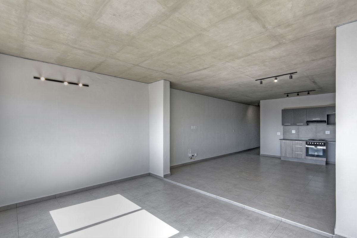 Spacious open-plan living area featuring an exposed concrete ceiling, modern tiled flooring, and a compact, functional kitchen with built-in appliances.
