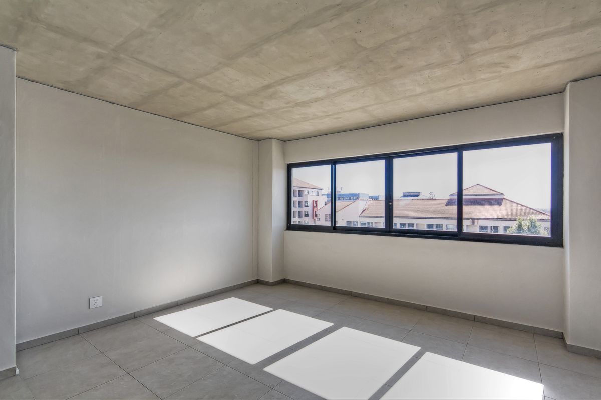 A bright, empty bedroom featuring an exposed concrete ceiling, neutral tiled flooring, and large aluminium-framed windows with urban views.