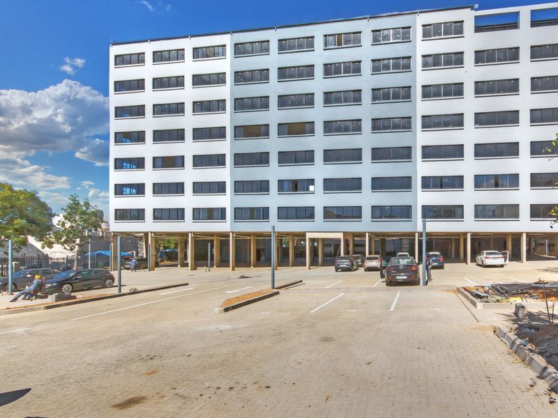 Exterior view of a multi-story apartment building featuring a paved ground-level parking area with covered bays and concrete structural columns.