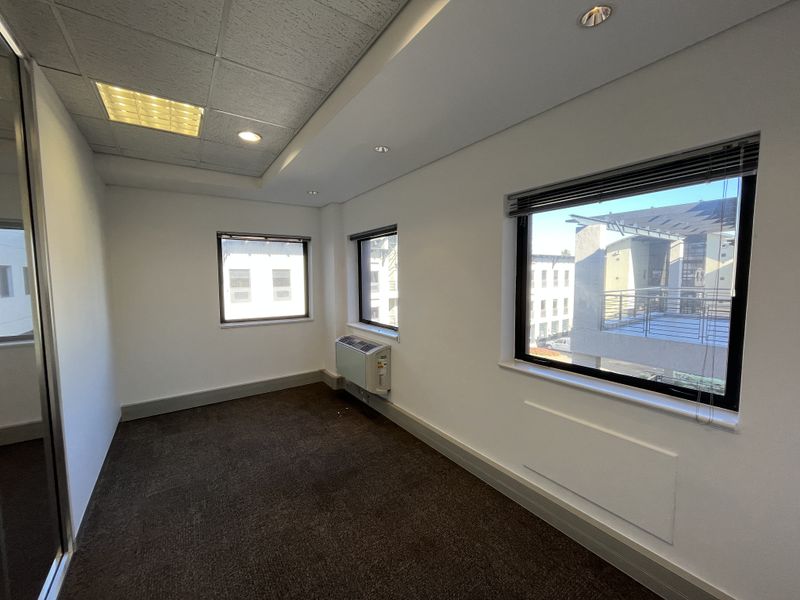 A bright, empty office space featuring carpeted flooring, recessed lighting, built-in cupboards, and an integrated air conditioning unit with large windows.