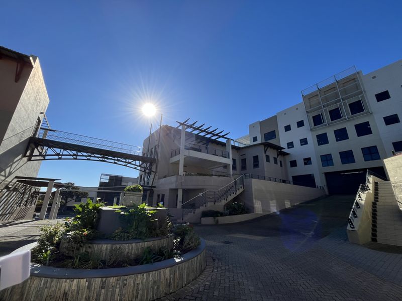Exterior view of a modern office complex featuring a pedestrian bridge, paved driveway, and landscaped stone planters under a clear blue sky.