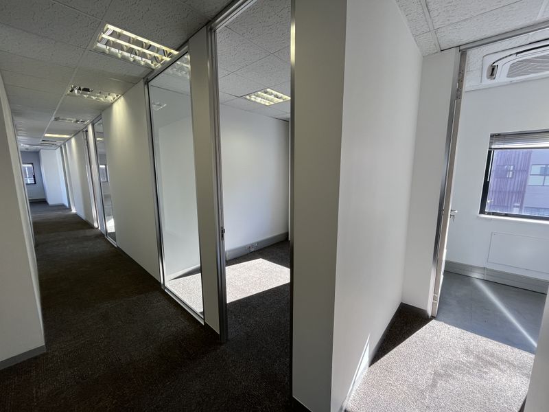 A bright, modern office corridor featuring glass partitions, suspended ceilings with recessed lighting, and carpeted flooring, providing a professional and clean workspace environment.
