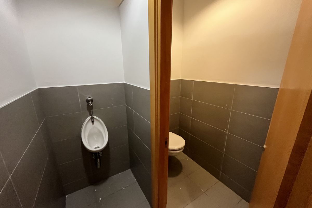 A functional office restroom featuring a wall-mounted urinal and a separate toilet stall with neutral grey wall and floor tiling.
