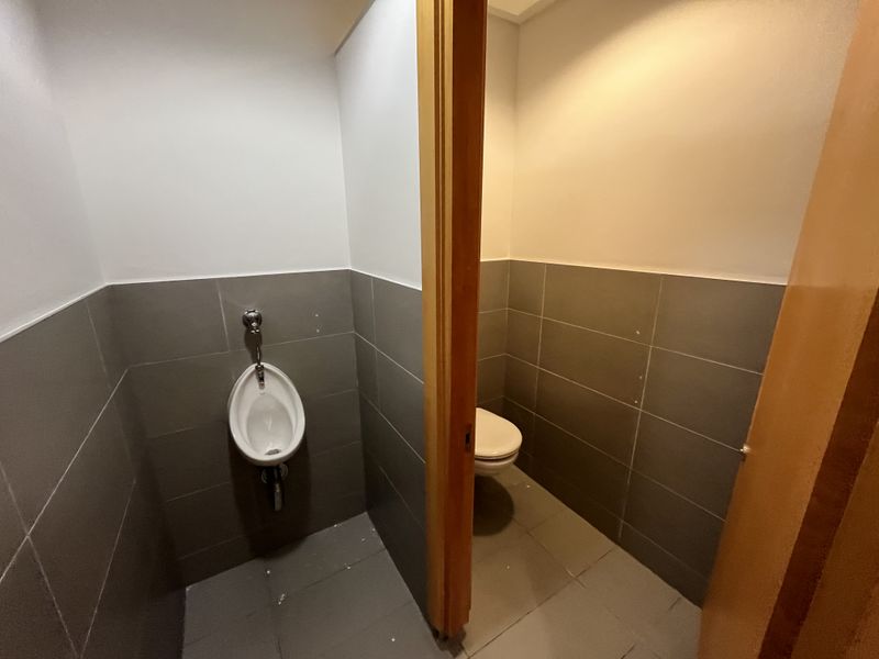 A functional office restroom featuring a wall-mounted urinal and a separate toilet stall with neutral grey wall and floor tiling.