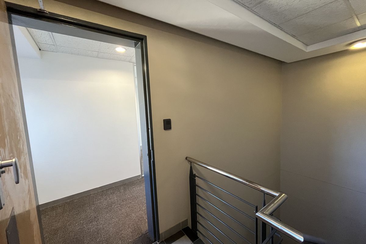 A clean office hallway featuring a stainless steel handrail, carpeted flooring, and recessed lighting leading into an adjacent office space.