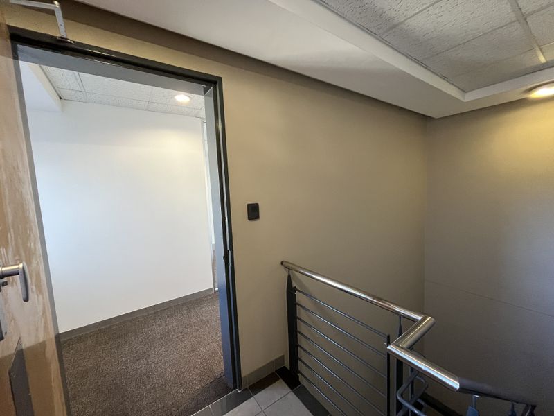 A clean office hallway featuring a stainless steel handrail, carpeted flooring, and recessed lighting leading into an adjacent office space.