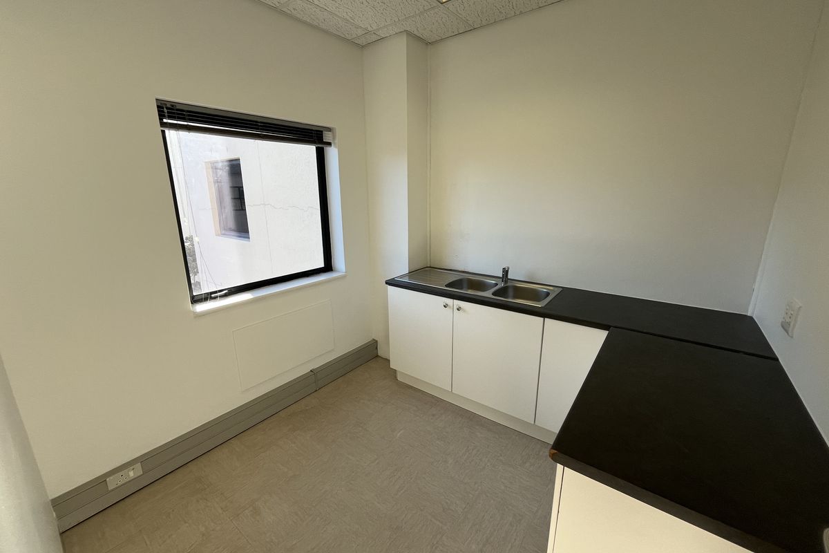 A clean, functional office kitchenette featuring built-in cabinetry, a stainless steel double sink, dark laminate countertops, and neutral vinyl flooring.