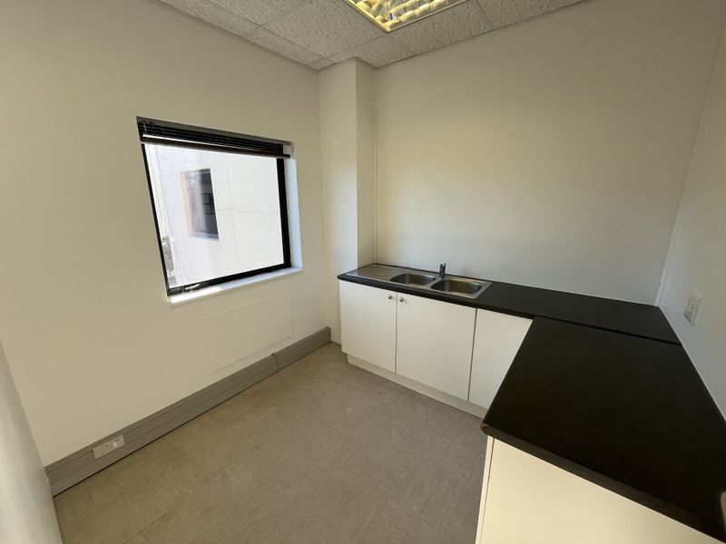 A clean, functional office kitchenette featuring built-in cabinetry, a stainless steel double sink, dark laminate countertops, and neutral vinyl flooring.