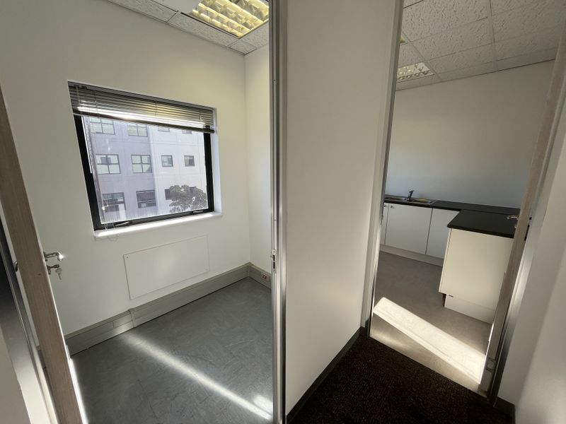 A bright, vacant office space featuring vinyl flooring, suspended ceilings, and an adjoining kitchenette area with built-in cabinetry and a sink.