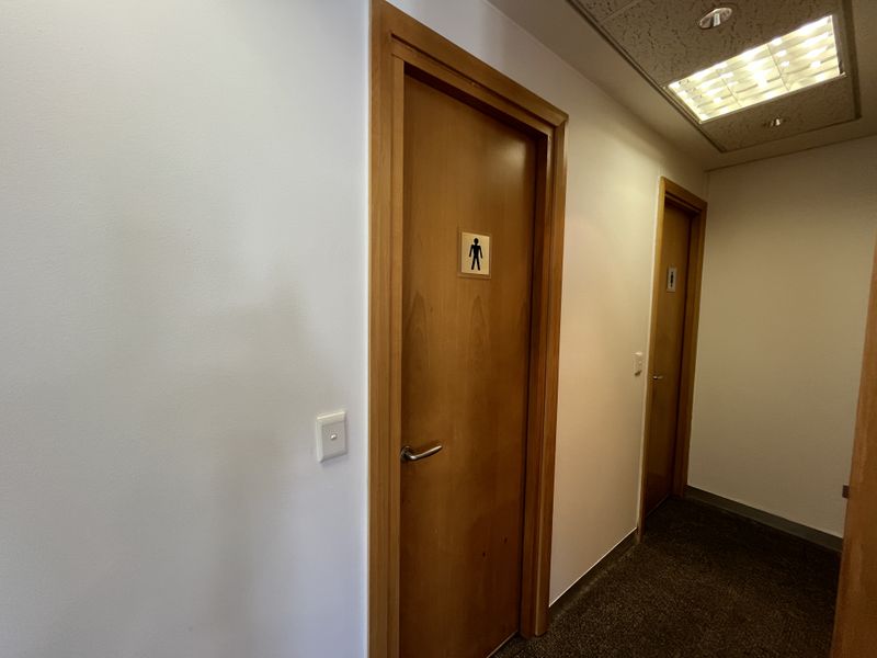 A functional office hallway featuring carpeted flooring, recessed ceiling lighting, and wooden doors leading to restroom facilities.