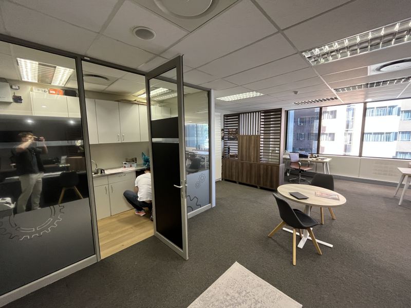 Modern office space featuring glass partitions, a small kitchenette area, suspended ceilings with recessed lighting, and large windows providing natural light.