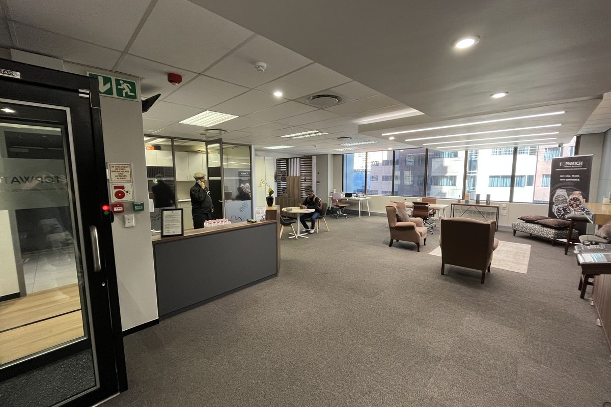 A spacious open-plan office area featuring suspended ceilings, recessed lighting, carpeted flooring, and large windows providing natural light.