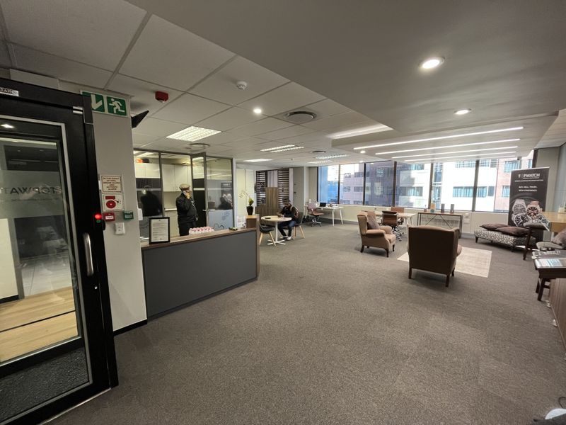 A spacious open-plan office area featuring suspended ceilings, recessed lighting, carpeted flooring, and large windows providing natural light.
