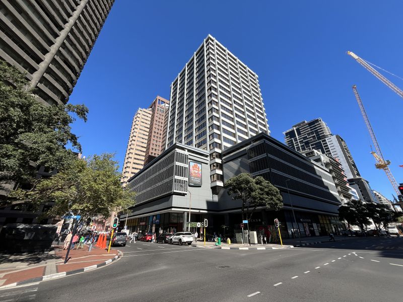 A wide-angle street view of a modern multi-story office building featuring a prominent glass facade and ground-level retail spaces in a busy urban environment.