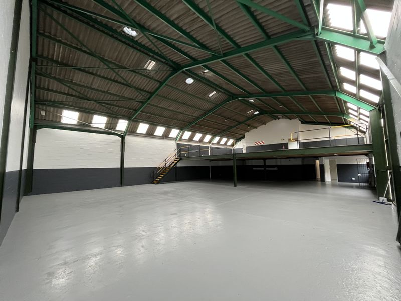 Spacious industrial warehouse unit featuring high ceilings with exposed steel trusses, integrated mezzanine level, ample natural light from skylights, and durable painted concrete flooring.
