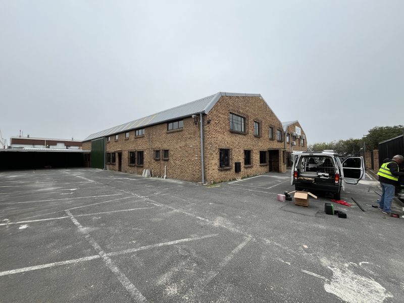 Exterior view of a face brick industrial building featuring a metal roof, security bars on windows, and an expansive asphalt parking area.