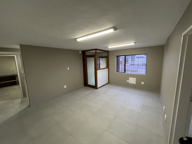 A clean, empty office space featuring neutral wall tones, tiled flooring, fluorescent lighting, and an internal glass partition with security bars on the window.
