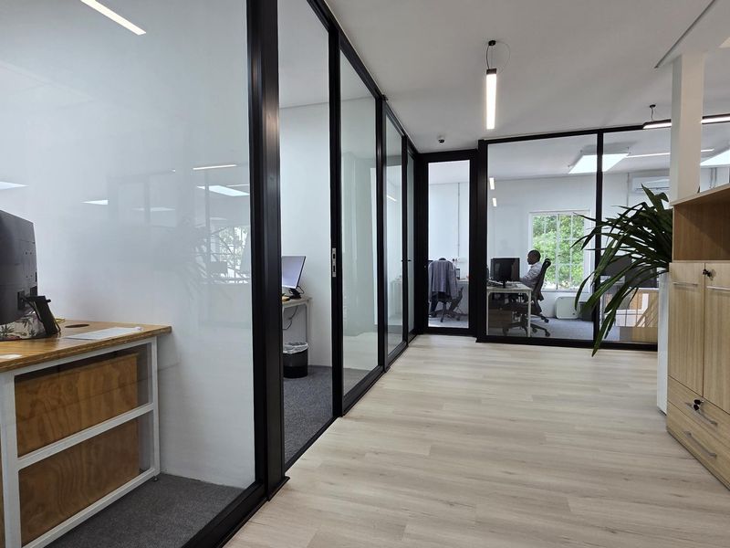 Modern office space featuring glass partition walls with black aluminum frames, light wood laminate flooring, and integrated storage cabinetry.
