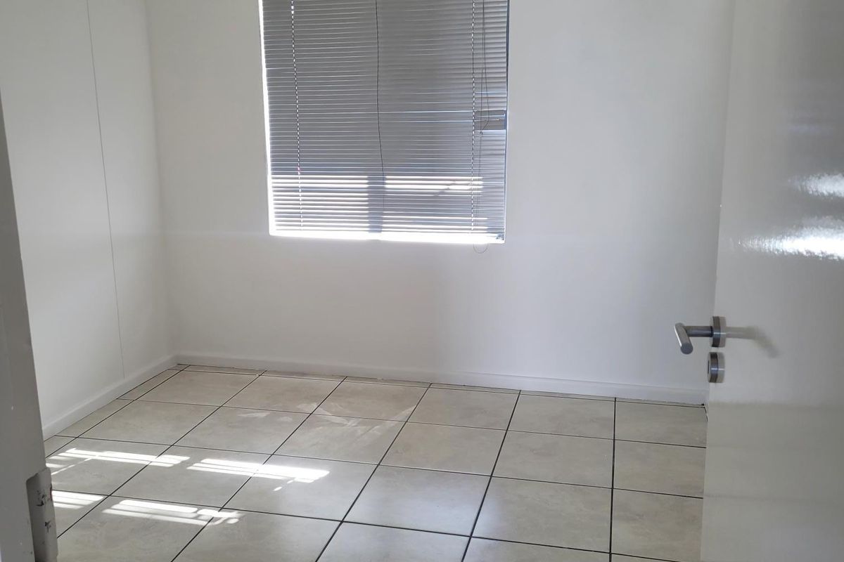 A clean, empty office space featuring neutral ceramic floor tiles, white walls, and a window fitted with venetian blinds.