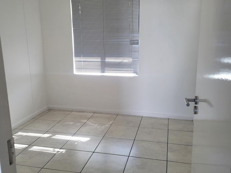 A clean, empty office space featuring neutral ceramic floor tiles, white walls, and a window fitted with venetian blinds.