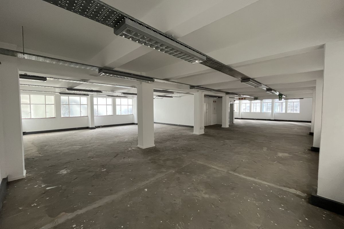 A large, open-plan office space featuring exposed cable trays, industrial fluorescent lighting, and unfinished concrete flooring, offering a blank canvas for customization.