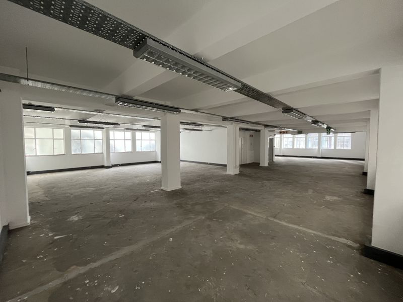 A large, open-plan office space featuring exposed cable trays, industrial fluorescent lighting, and unfinished concrete flooring, offering a blank canvas for customization.