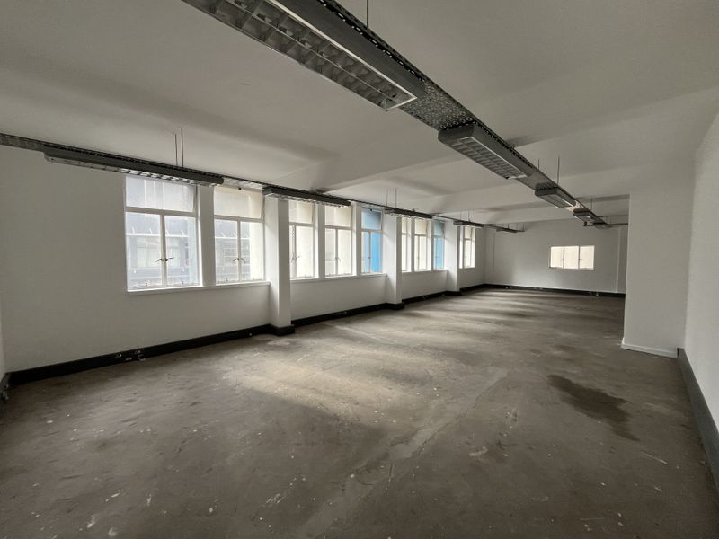 A spacious, empty open-plan office area featuring industrial-style suspended lighting, exposed cable trays, and large windows providing ample natural light.