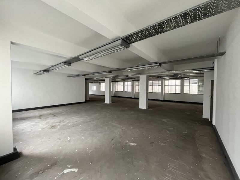 A large, open-plan office space featuring exposed cable trays, industrial lighting, concrete flooring, and expansive windows providing natural light.