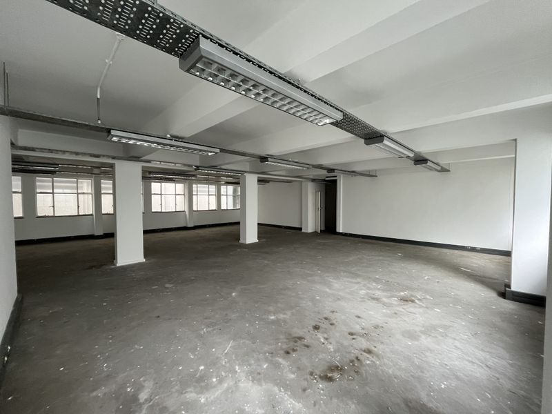 Spacious open-plan office area featuring exposed ceilings, industrial lighting, concrete flooring, and large windows providing ample natural light.