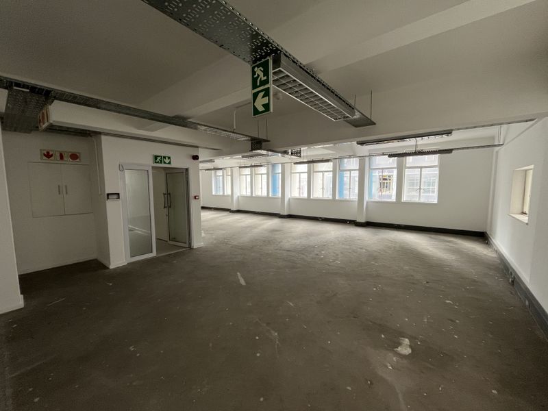 An open-plan office space featuring exposed cable trays, industrial lighting, and large windows, currently in a shell condition with unfinished flooring.