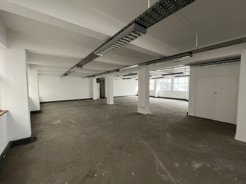 Spacious open-plan office area featuring exposed cable trays, industrial-style lighting, structural columns, and concrete flooring, offering a versatile blank canvas for business operations.