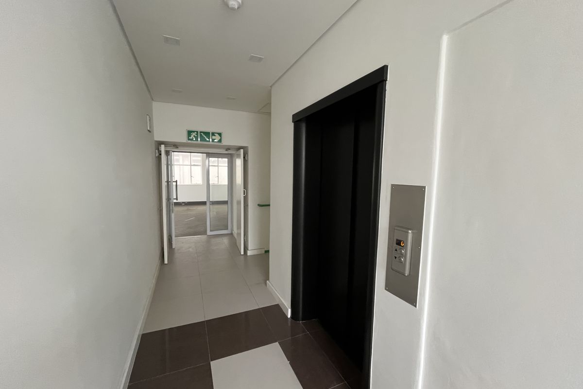 A clean, modern office hallway featuring tiled flooring, a built-in elevator, and clear fire exit signage leading to an adjacent office space.