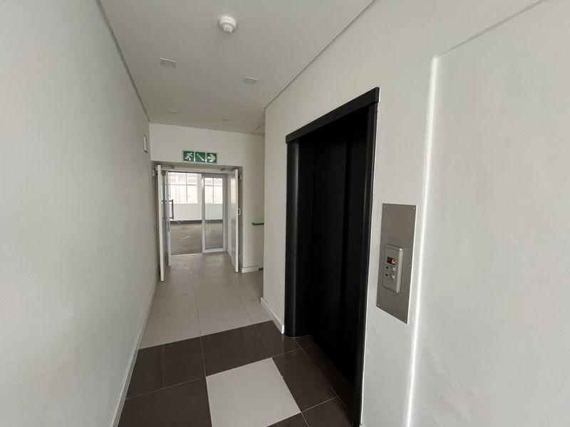 A clean, modern office hallway featuring tiled flooring, a built-in elevator, and clear fire exit signage leading to an adjacent office space.