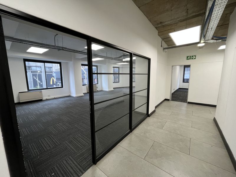 Modern open-plan office space featuring glass partitions, carpet tiles, and an exposed concrete ceiling with recessed lighting.