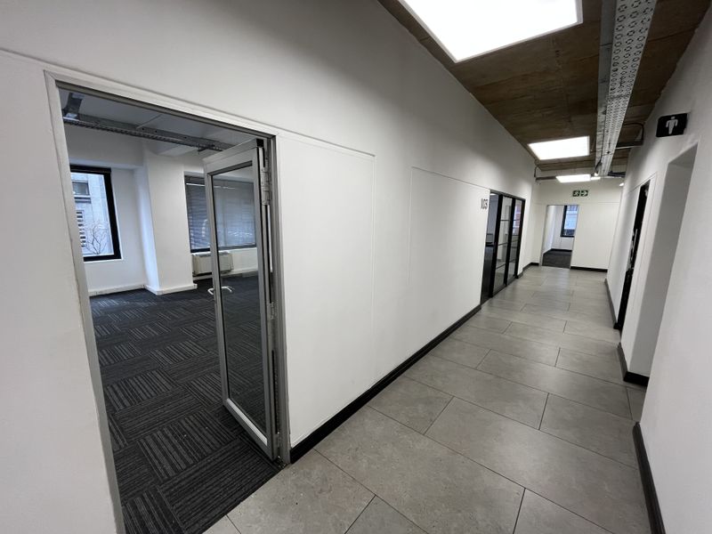 A modern office hallway featuring tiled floors, exposed concrete ceilings with recessed lighting, and glass-paneled doors leading into vacant office spaces.