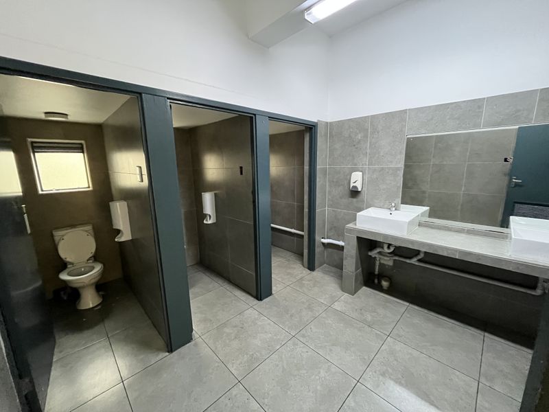 A functional office bathroom featuring tiled finishes, multiple toilet cubicles, and a built-in vanity with dual basins.