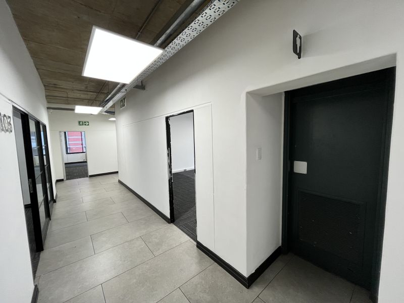 A modern office hallway featuring tiled flooring, exposed concrete ceilings with LED panel lighting, and access to multiple office suites and restroom facilities.