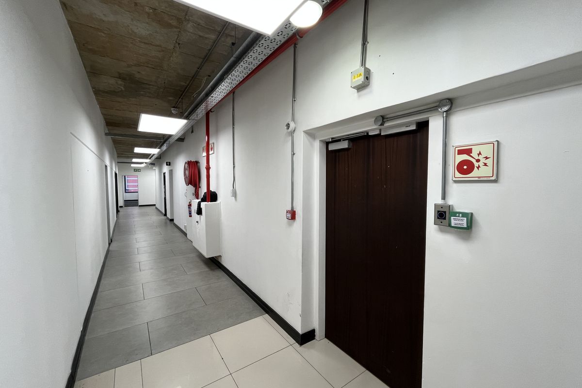 A clean, modern office hallway featuring exposed concrete ceilings, tiled flooring, and integrated fire safety infrastructure including a hose reel and alarm system.