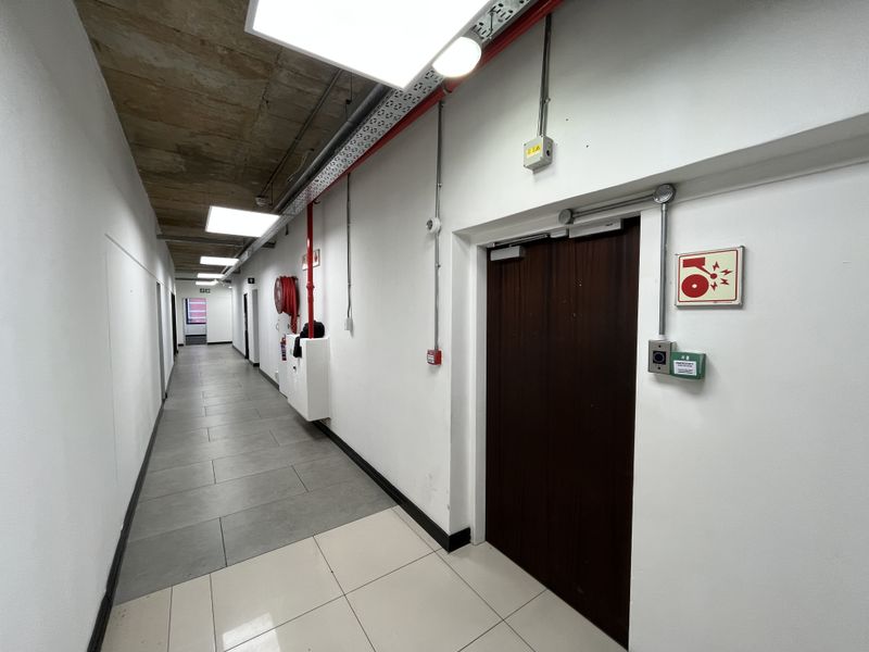 A clean, modern office hallway featuring exposed concrete ceilings, tiled flooring, and integrated fire safety infrastructure including a hose reel and alarm system.