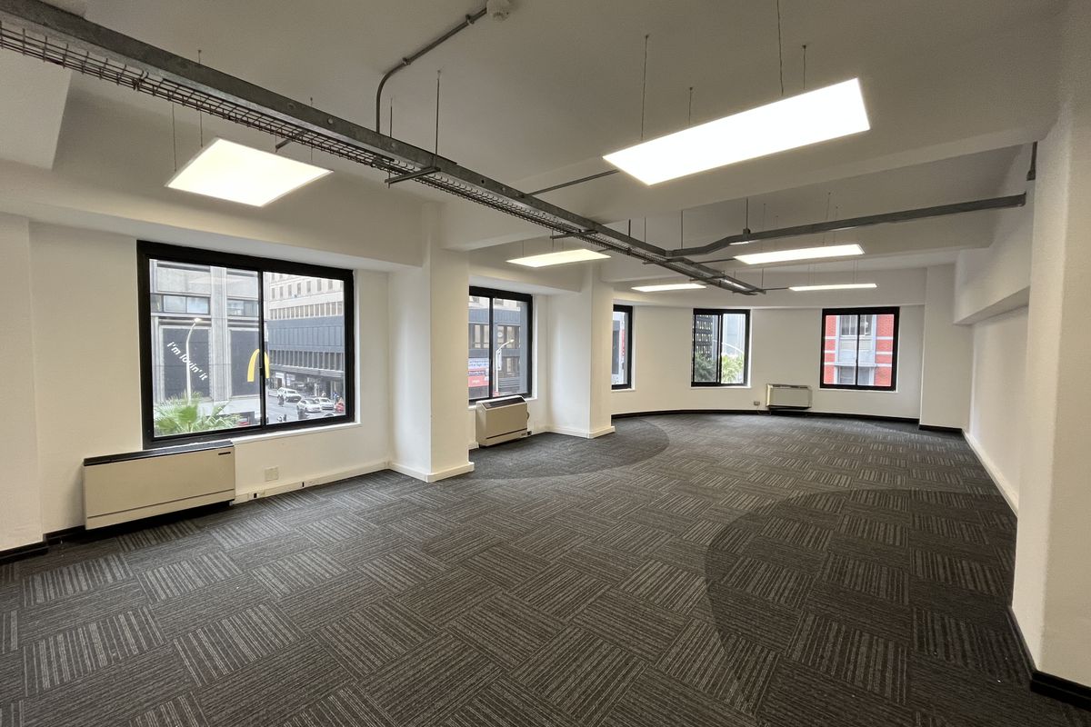 Spacious open-plan office area featuring carpet tiles, suspended LED lighting, exposed cable management, and multiple windows providing natural light and city views.