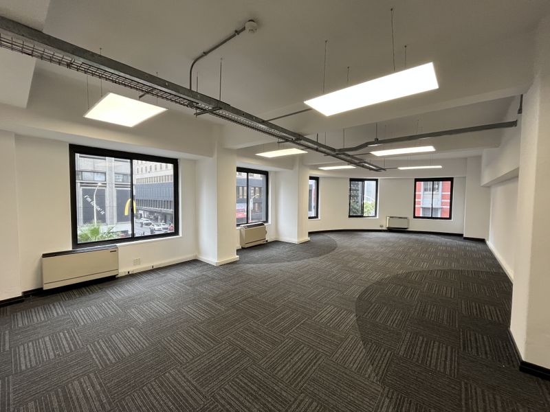 Spacious open-plan office area featuring carpet tiles, suspended LED lighting, exposed cable management, and multiple windows providing natural light and city views.