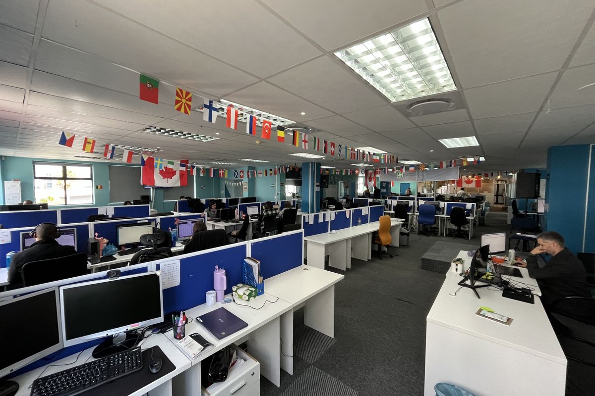 A large open-plan office space featuring rows of built-in workstations, suspended ceilings with recessed lighting, and carpeted flooring throughout.