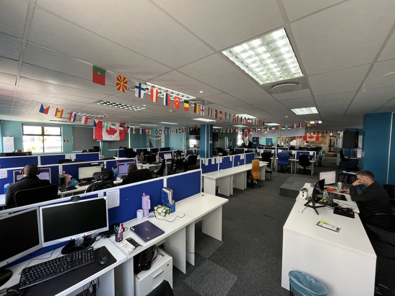 A large open-plan office space featuring rows of built-in workstations, suspended ceilings with recessed lighting, and carpeted flooring throughout.