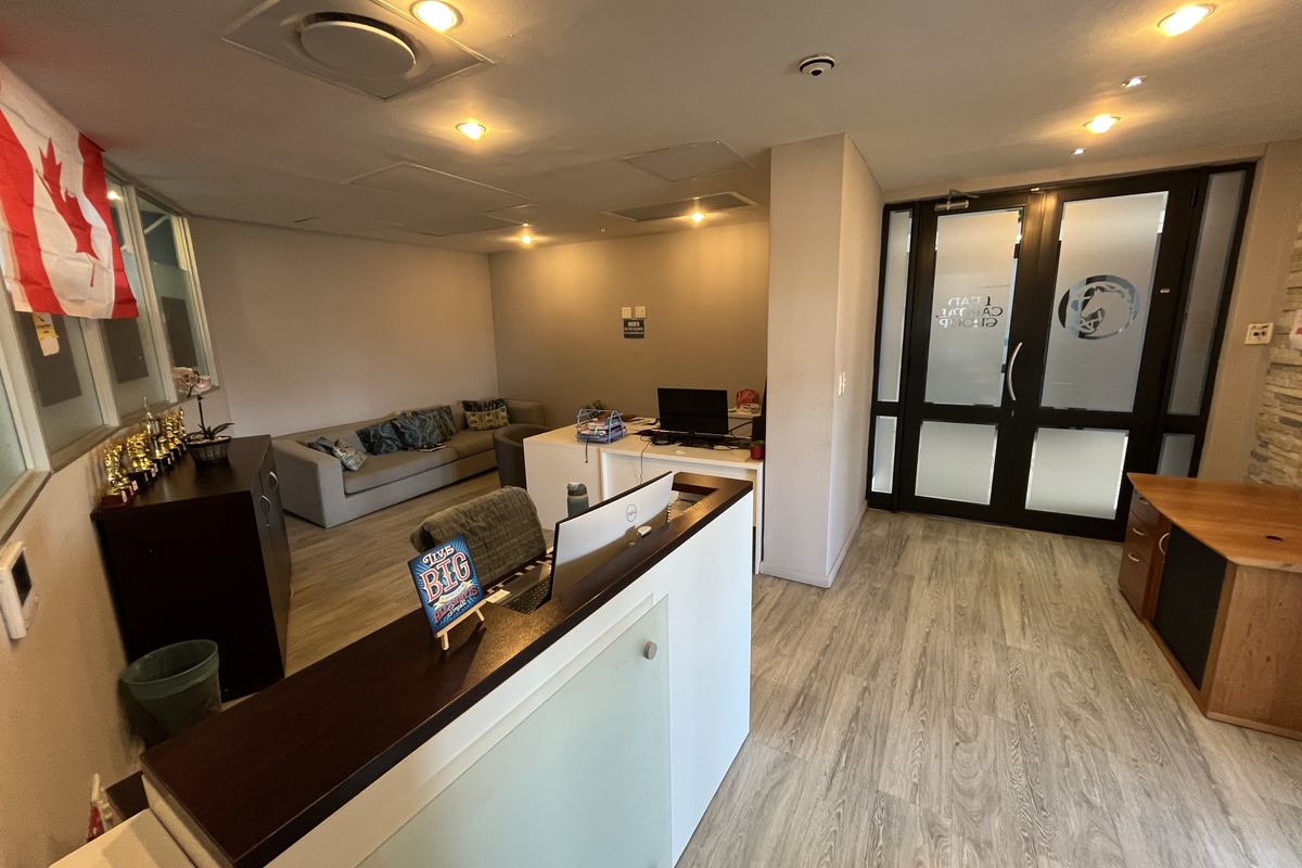 A professional office reception area featuring a modern reception desk, laminate flooring, recessed lighting, and glass-paneled double entry doors.