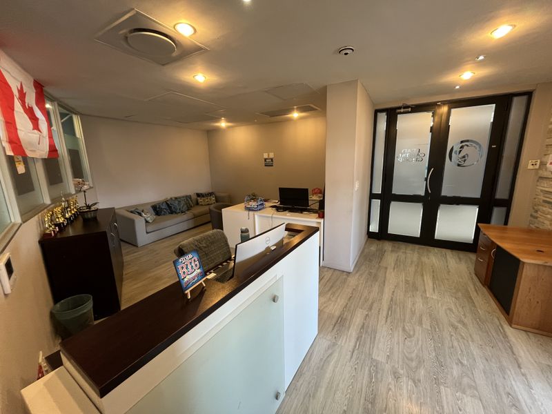 A professional office reception area featuring a modern reception desk, laminate flooring, recessed lighting, and glass-paneled double entry doors.