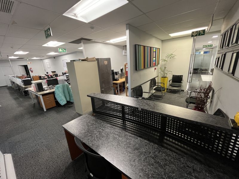 Open-plan office space featuring a reception desk, partitioned work areas, suspended ceiling with recessed lighting, and carpeted flooring throughout.