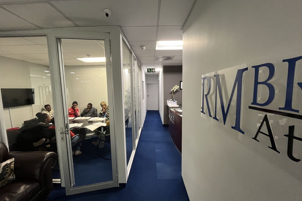 A professional office hallway featuring glass-partitioned meeting rooms, suspended ceilings with recessed lighting, and blue carpeting throughout the workspace.